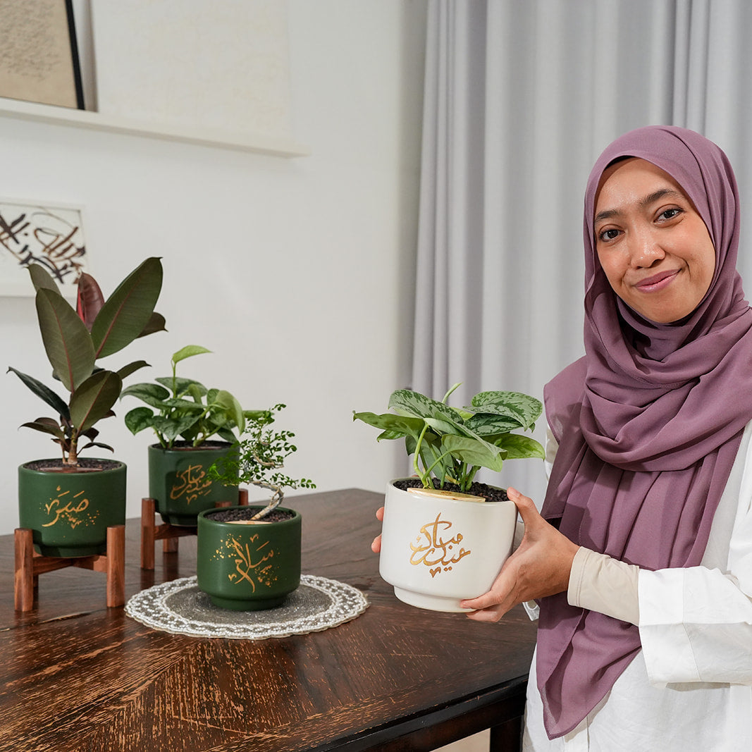 Shatila holding a potted plant in a home setting with other plants on a table.